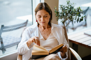 White mature woman smiling and reading book while sitting in cafe