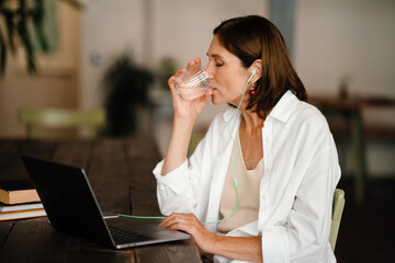 White mature woman drinking water and working with laptop in cafe