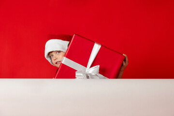 Boy in Santa's holiday hat hides behind a large red christmas birthday gift box. Preschool cute...