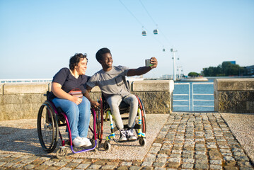 Happy biracial couple taking selfie on embankment. African American man and Caucasian woman in wheelchairs on embankment, holding mobile phone, smiling. Love, relationship, social media concept - Powered by Adobe