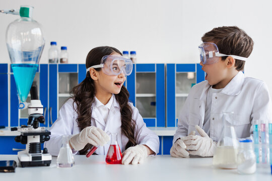 amazed girl in goggles looking at friend near flask with red liquid in chemical lab.