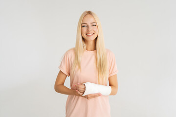 Studio portrait of cheerful blonde young woman with broken arm wrapped in plaster bandage smiling looking at camera, standing on white isolated background. Concept of insurance and healthcare. - obrazy, fototapety, plakaty