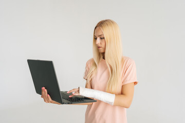 Portrait of serious pretty young woman with broken arm wrapped in plaster bandage holding laptop and working typing, standing on white isolated background. Concept of insurance and healthcare.