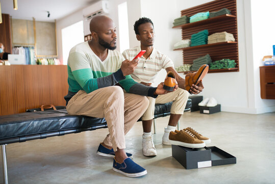Portrait of African gay couple taking photo of modern shoes. Two handsome men sitting on bench choosing pair of shoes to buy using phone for taking picture. LGBT couples leisure, buying shoes concept