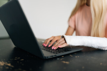 Close-up side view of unrecognizable young woman with broken hand wrapped in plaster bandage typing working on laptop computer, sitting at black table on white isolated background.