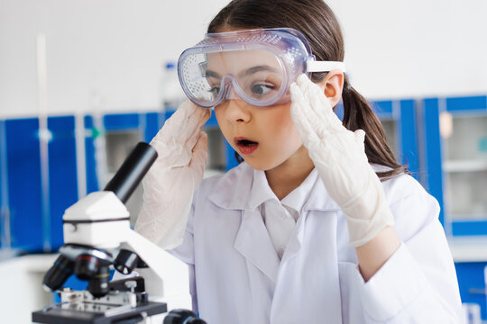 shocked girl in goggles and latex gloves touching head near microscope in laboratory.