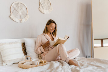 pretty smiling woman relaxing at home on bed in morning in pajamas reading book - Powered by Adobe