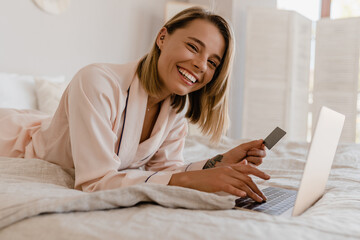 pretty smiling woman shopping online on laptop at home on bed in morning in pajamas