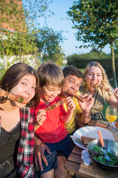 Hungry family sitting around table. Mid adult parents and children looking at camera, eating meat canapes and steak, making faces. BBQ, cooking, food, family concept
