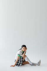Trendy asian model in cropped cardigan looking at camera while sitting on grey background