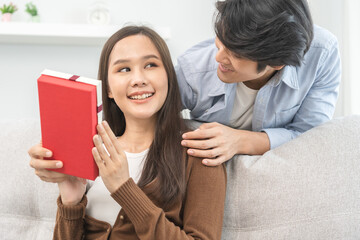 Celebrating Valentine's day anniversary, relationship two asian young couple love, boyfriend gives a gift to girlfriend by hide box at the behind, woman getting present while sitting on sofa at home.