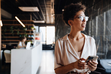 Happy black businesswoman smiling and using cellphone in office lobby