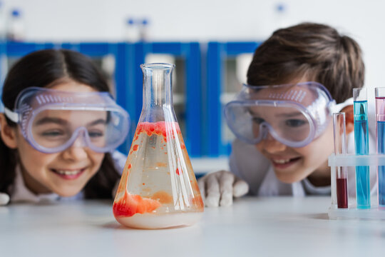 excited kids in goggles looking at flask with liquid and red substance in chemistry lab.