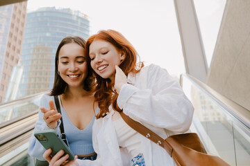 Young multinational women using cellphone while standing on escalator