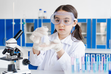 girl in goggles holding flask near microscope in laboratory.
