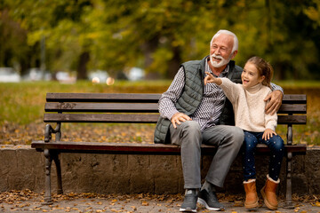 Grandfather spending time with his granddaughter on bench in park on autumn day - obrazy, fototapety, plakaty