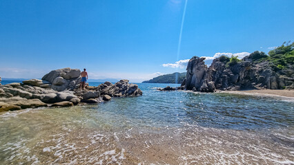 Man in swimsuit staning on rocks with panoramic view of holy Mount Athos from Fava sand beach,...