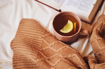Winter aesthetic flat lay photo. A cup of hot tea with lemon, open book and brown knitted sweater on a bed. Weekend morning
