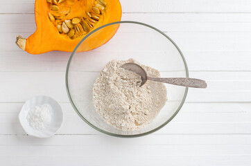 Glass bowl with oatmeal flour, half a pumpkin with seeds and baking powder on a white wooden background. Cooking healthy vegan food