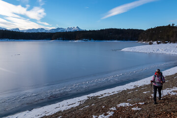 Woman with hiking backpack walking along the shore of the frozen lake Forstsee, Techelsberg, Carinthia (Kaernten), Austria, Europe. Winter wonderland and snow landscape. View of Karawanks mountains