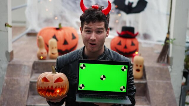 Handsome Young man dressed as a vampire devil with makeup holds a pumpkin Jack O Lantern and modern laptop with green screen mockup with chroma key with tracking dots