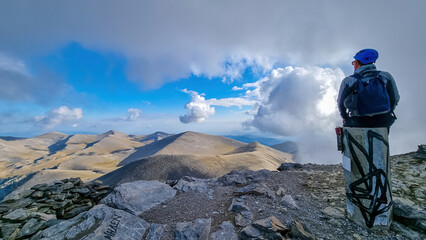 Man with helmet sitting on cloud covered mountain summit of Skolio peak on Mount Olympus, Mt Olympus National Park, Macedonia, Greece, Europe. View of rocky ridges and highlands from throne of Zeus