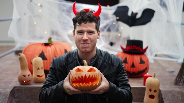 Handsome Young man dressed as a vampire devil with makeup holds a pumpkin Jack O Lantern. Halloween props, bats, pumpkins, skulls, spiders, brooms, burning candles, cobwebs. The concept of application