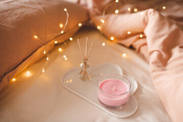 Cozy home atmosphere with pink scented candle on white ceramic tray and liquid diffuser in bed over...