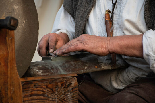 close up craftmanship, man sharpening knife on whetstone. Traditional handwork, historic outfit. Middle aged white male at work in workshop. Detail hands. 