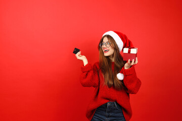 Happy woman consumer in a santa hat and a credit card in her hands on a red background. New Year's...