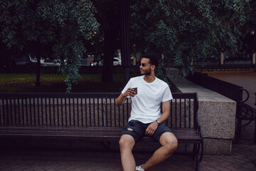 African american man in a white t-shirt sits on a bench and drinks coffee. Mock-up.