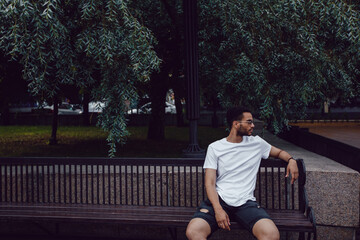 Fototapeta premium African american man in a white t-shirt sits on a bench in the street. Mock-up.
