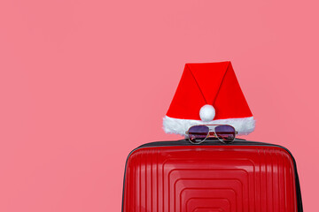 Close-up of a red suitcase with a red Santa hat and sunglasses, isolate on a pink background. The concept of traveling during the Christmas holidays.
