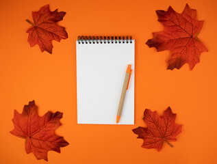 Naklejka na ściany i meble Autumn composition with blank notepad, pen and autumn leaves on orange background. Workplace. Flatlay. Top view. Copy space.