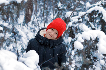 A happy smiling boy in a black down jacket and a red knitted hat plays and runs in a beautiful snowy winter park for Christmas