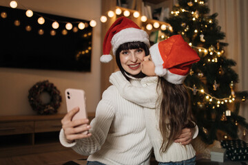 Fototapeta premium Little cute preschool girl daughter kissing her smiling caucasian female mother. Mom taking selfie with small female kid in santa hat, sitting on floor on background of decorated Christmas tree.