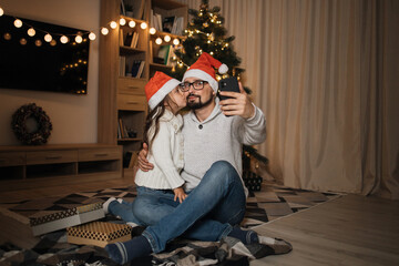 Little cute preschool girl daughter kissing her smiling caucasian male father. Daddy taking selfie with small female kid in santa hat, sitting on floor on background of decorated Christmas tree.