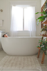 Woman peacefully soaking in deep white tub in bright light-filled bathroom with houseplants