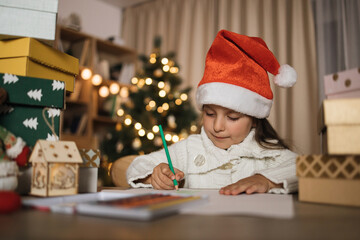 Letter for Santa. Cute little caucasian girl in red hat sitting at table and writing wish list of presents for Christmas in decorated room.