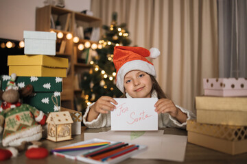 Christmas, holidays and childhood concept - smiling caucasian girl in red hat holding letter in hands after writing wish list for Santa at home.