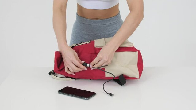 Young woman with beautiful figure, an open belly in gray leggings connects her phone to USB charging port in red backpack, 4k