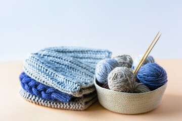 Gray, brown and blue balls of knitting threads in a beige basket with bamboo knitting needles and a stack of knitted hats for winter and autumn, knitting hobby