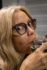Blonde woman with glasses and crazy look drinking from two glasses of wine simultaneously.