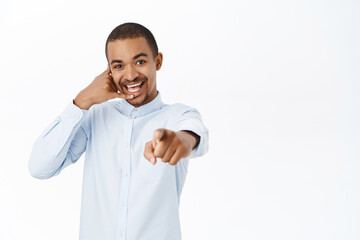 Smiling guy shows telephone gesture, points at camera, call us gesture, call center advertisement, standing over white background