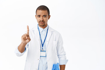 Fototapeta premium Serious doctor shows stop gesture, one finger, talking to patient, standing concerned over white background