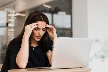 Woman manager with strong headache working on laptop online remotely in white office. Sad girl with laptop got migraine touches her head because of pain.