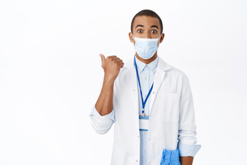 Surprised doctor in medical mask, points left, shows interesting advertisement, clinic promo offer, stands over white background