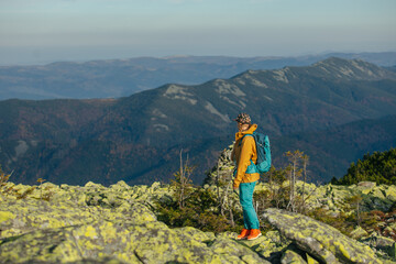 a girl hiker in the mountains with a blue backpack walks against the background of incredibly beautiful mountains. A girl in a cap and a mustard-colored jacket walks along the rocks