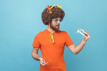 Portrait of funny man with Afro hairstyle wearing orange T-shirt choosing colorful eyeglasses,...