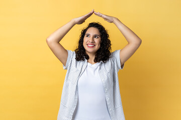 I'm in safety. Portrait of woman with dark wavy hair raising hands showing roof gesture and smiling contentedly, dreaming of house, looking away. Indoor studio shot isolated on yellow background.